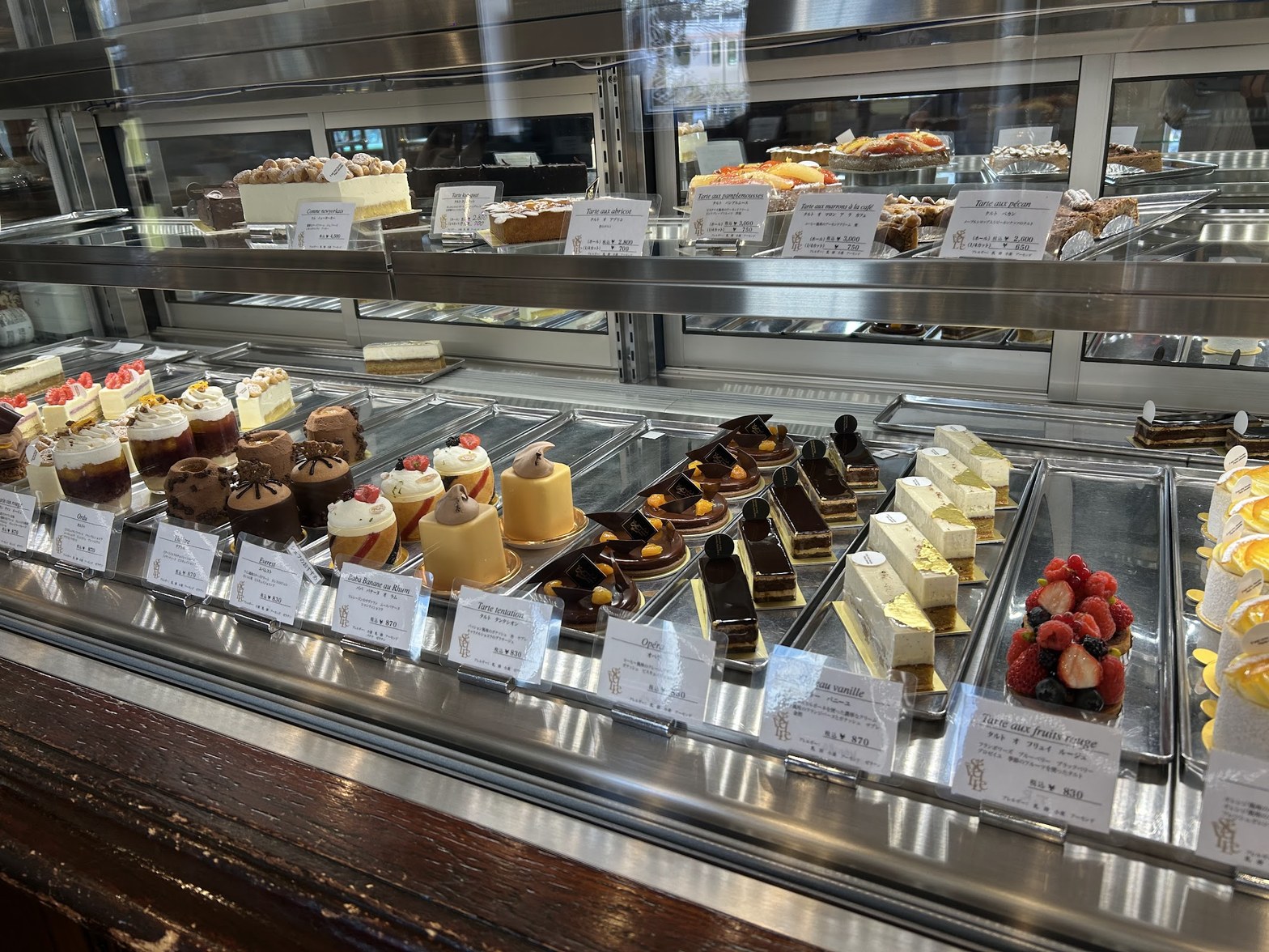 ガラスショーケースにケーキ・タルト・チョコレート菓子など20種以上がプライスカードとともに2段に並ぶ店内の様子