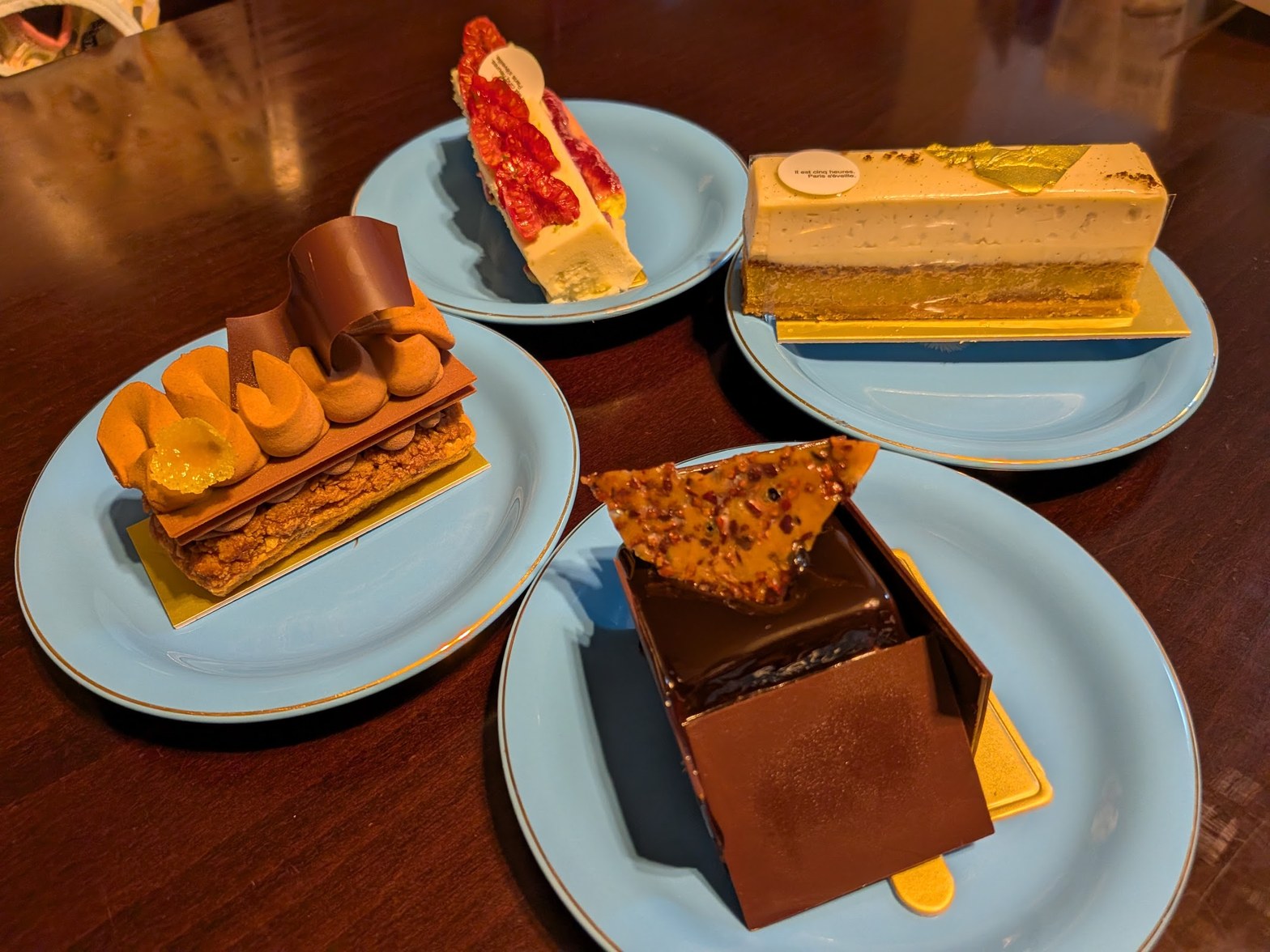 暗い木のテーブルに青い丸皿4枚が並び、ミルフィーユ・チョコレートケーキ・フルーツタルト・バニラムース風のケーキが盛り付けられている