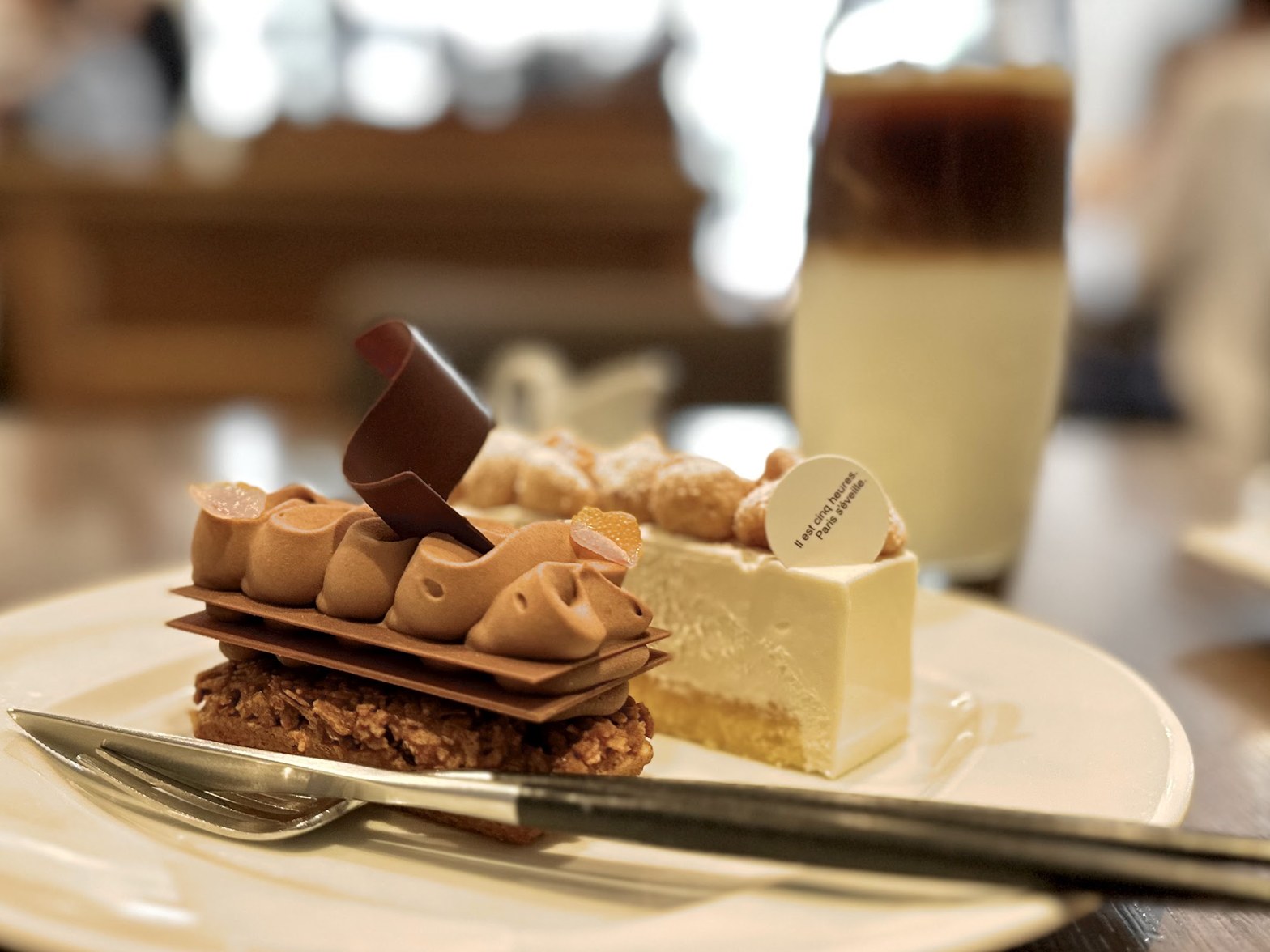 白い大皿にチョコレート系ミルフィーユとバニラ系ケーキが並び、奥にアイスコーヒーの入ったグラスが見える